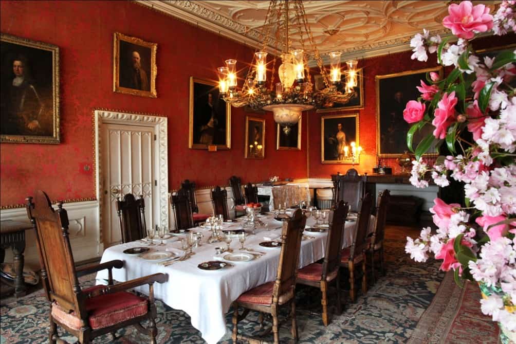 An ornate dining room with a long table covered by a white cloth and set with tableware. The room has red walls with many framed portraits, and a chandelier over the table.